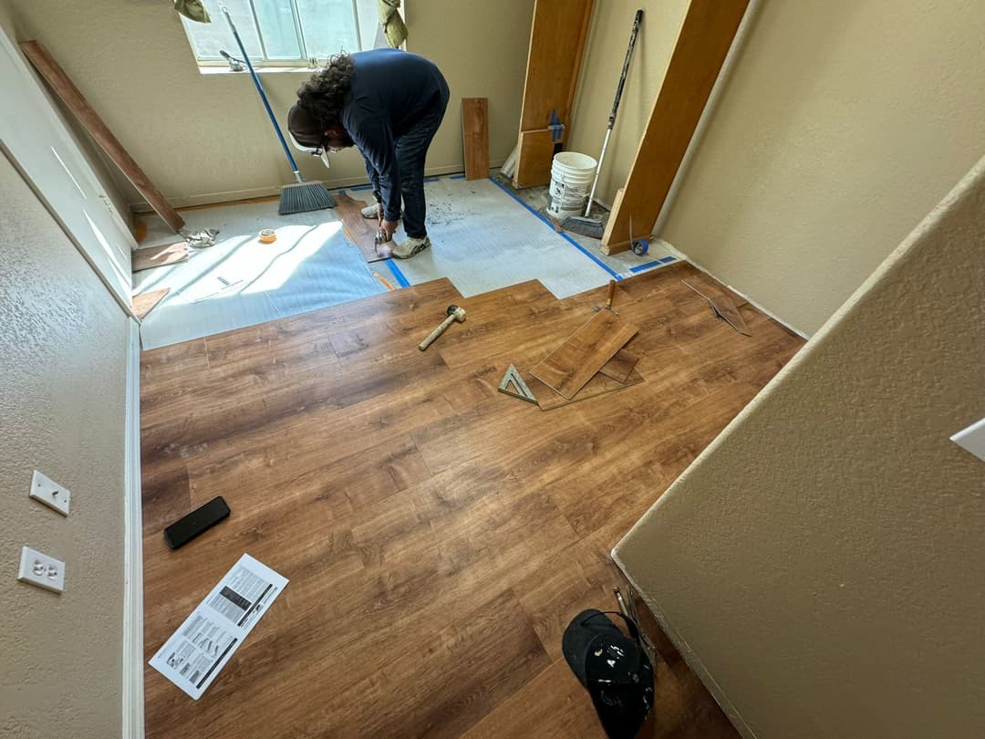 Person installing laminate flooring in a room with wood planks and tools on the floor.