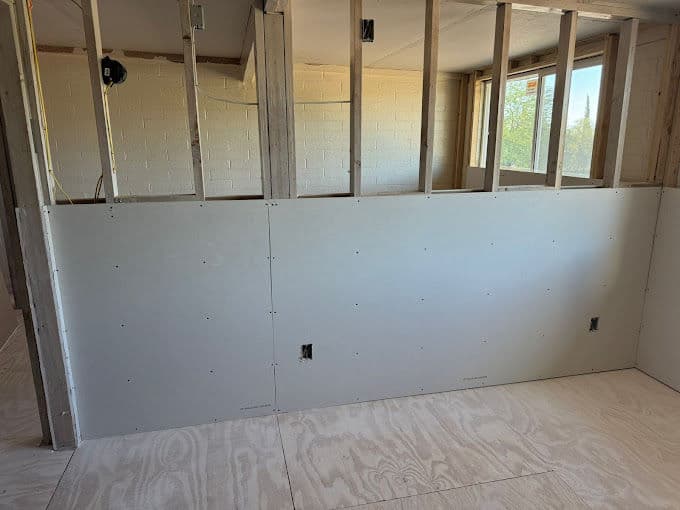 Drywall installation in a framed room with unfinished walls and plywood flooring.
