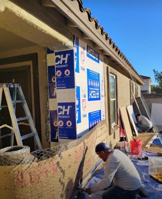 Worker applying exterior insulation to a house under construction with ladders and materials nearby.
