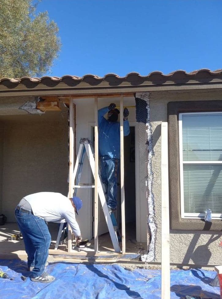 Construction workers renovating a doorway with ladders and tools on a sunny day.
