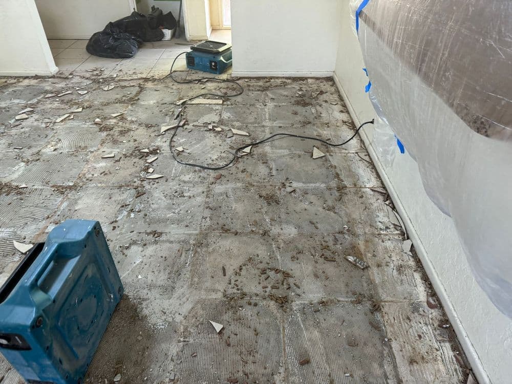 Renovation site with debris, exposed floor, blue tools, and plastic sheeting.