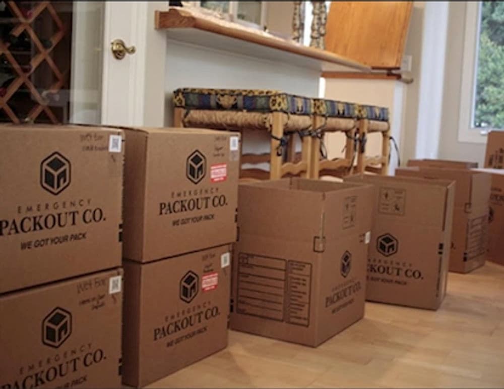 Cardboard moving boxes stacked in a home's entryway, labeled 'Emergency Packout Co.'